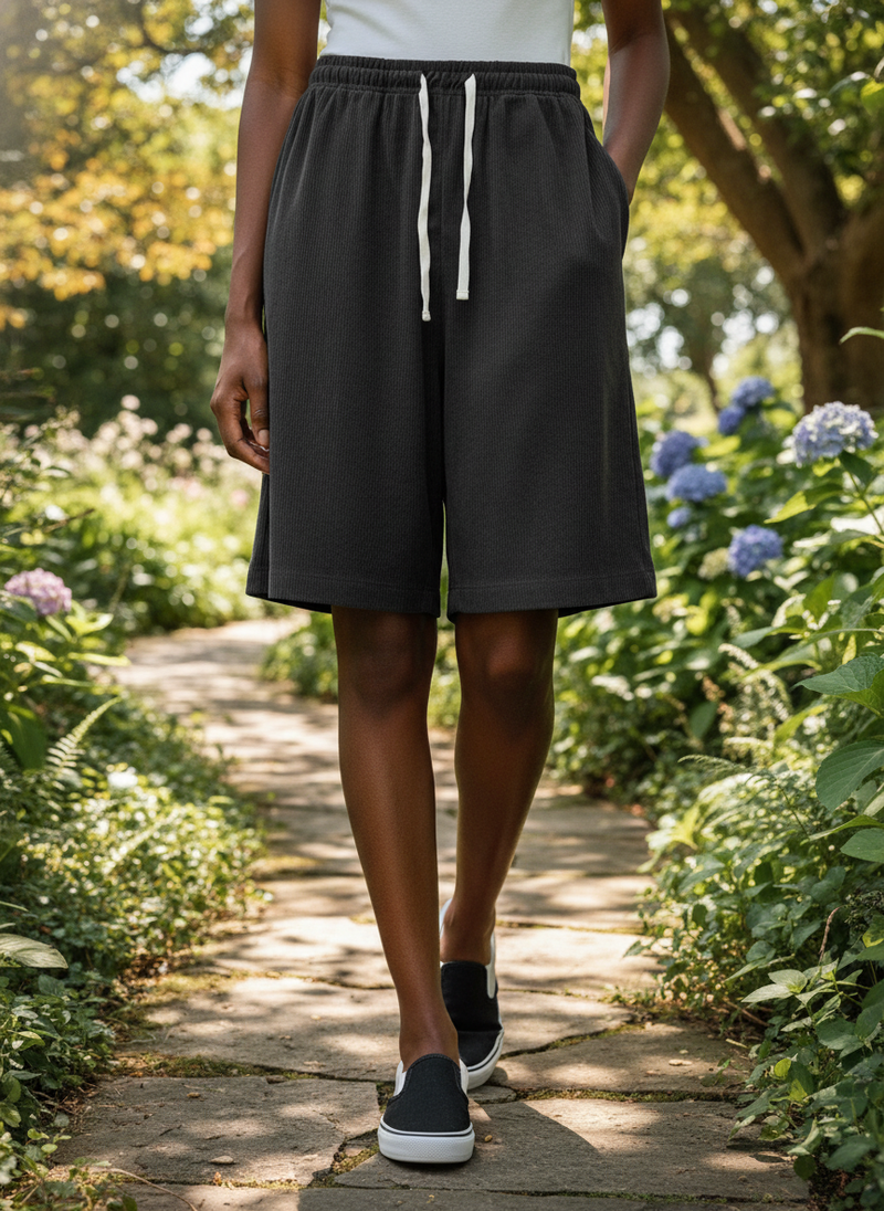 A person walks through a sun-dappled garden path, wearing relaxed, wide-leg CORDMERE corduroy shorts with a ribbed texture.